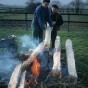 Zwei Personen legen grob zugerichtete Holzbalken mit ihrer Spitze in ein Feuer.