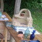 Hinter übereck stehenden Holztischen sitzt ein römisch gewandeter Kollege. Auf den Tischen steht das Modell eines Glasofens und verschiedene blaue Glasgefäße, daneben liegen Zangen und Informationsbroschüren.