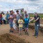 Eine Gruppe Besucher steht vor einer Eintiefung in der Erde, eine Archäologin deutet mit den Händen zur Vertiefung.