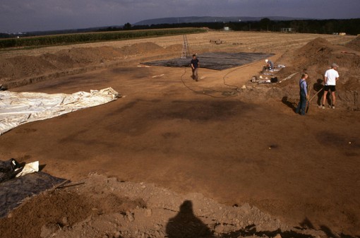 Photo der Ausgrabungfläche, im Hintergrund die Sophienhöhe, im Vordergrund schwarzbraune Verfärbung im hellbraunen Boden. Drei Personen, teilweise mit Schaufel, arbeiten auf der Fläche.
