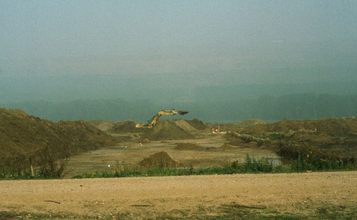 Das Foto zeigt die Grabungsfläche des Fundplatzes unmittelbar hinter einem mit Kies befestigten Weg. Im Hintergrund legt ein Bagger die Grabungsfläche weiter frei. Erdhügel auf der Fläche markieren die Stellen, wo Überreste der Siedlung ausgegraben werden.