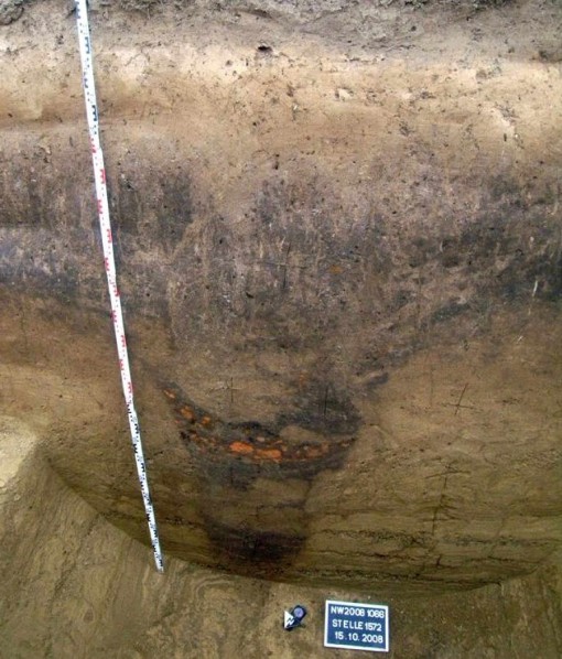 Foto: Profil einer über 2 Meter tiefen, neolithischen Schlitzgrube des Fundortes Merzenich. Man erkennt in der Profilgrube eine Verfüllung mit dunkel gefärbtem Oberbodenmaterial, das sich deutlich vom umgebenden, hellen Bodenmaterial abgrenzt. Man geht davon aus, dass dieses dunkle Material dem prähistorischen, alten Ackerboden entspricht.