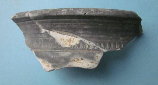 Das Foto zeigt die Fragmente einer spätantiken, sogenannten Fußschale aus oxidierend gebranntem Ton. Die Ware wird als Terra Nigra bezeichnet und weist eine hell- bis dunkelgraue Oberfläche auf, während das Innere der Scherben weiß bis gelblich ist.